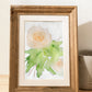 An art print of a peach rose painted with watercolors in a loose, impressionist style inside of a wooden frame on a table next to a candle.