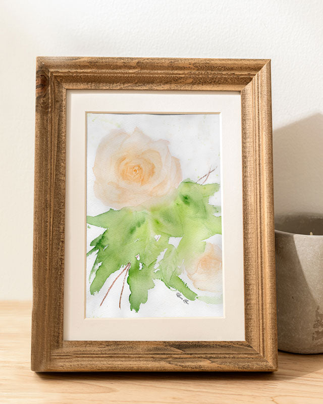 An art print of a peach rose painted with watercolors in a loose, impressionist style inside of a wooden frame on a table next to a candle.