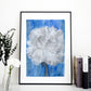 Art print of a mix media abstract floral painting of a white carnation on a blue background in a black frame on a shelf next to books and vases of flowers.