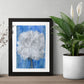 Art print of a  mix media abstract floral painting of a white carnation on a blue background in a black frame on a table in between two plants.